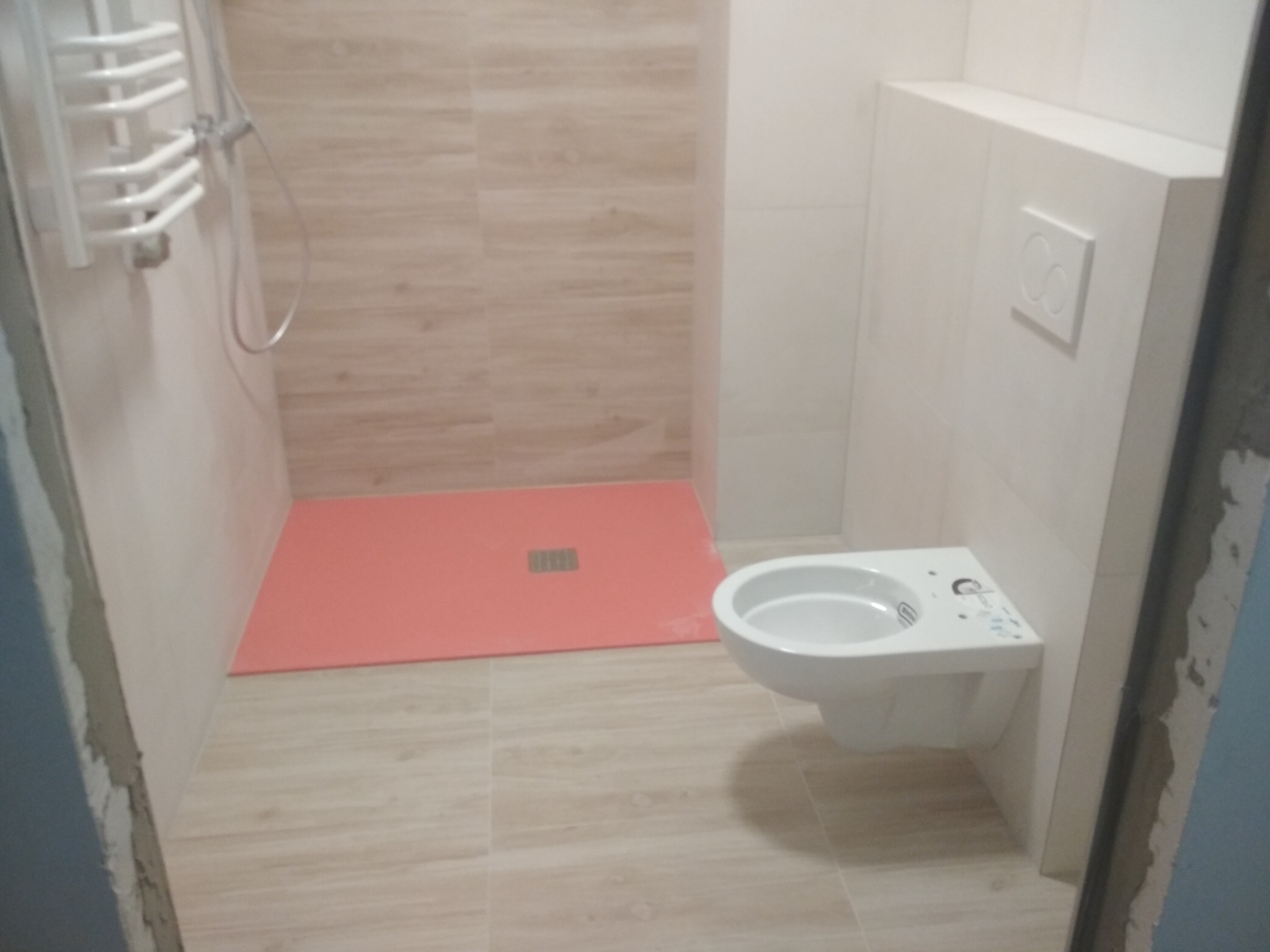 Świeżo wykończona łazienka z beżowymi płytkami imitującymi drewno, z zamontowaną podwieszaną toaletą i brodzikiem z odpływem liniowym w kolorze koralowym.