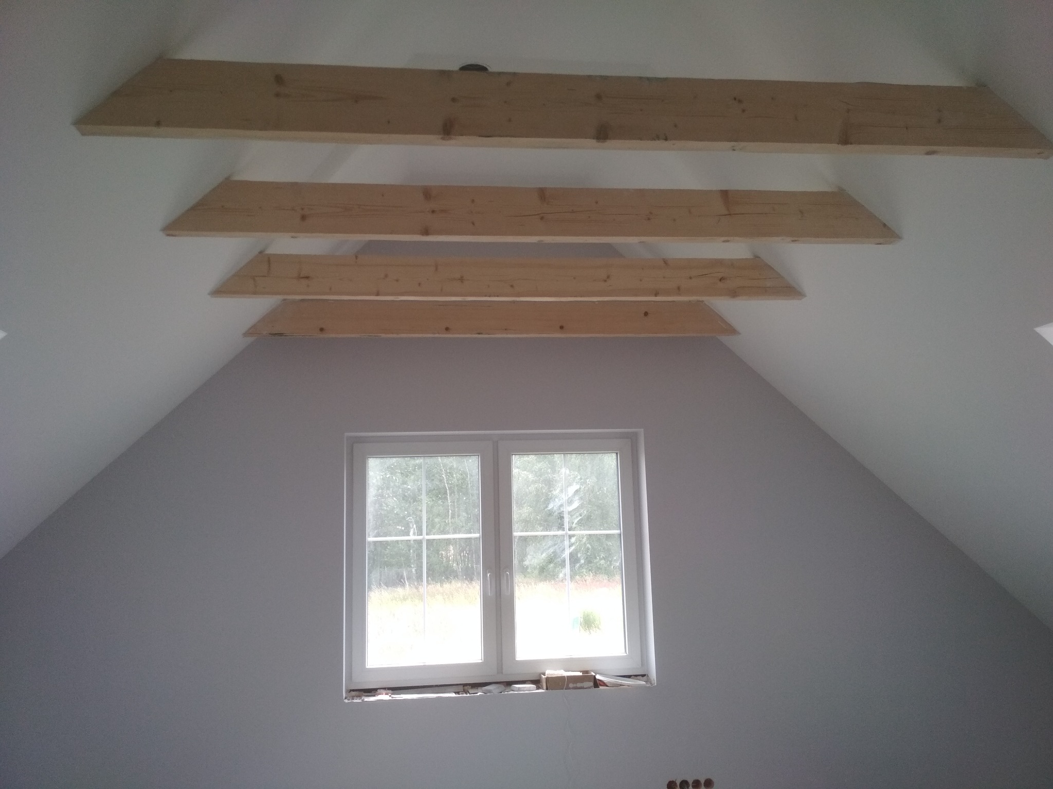 Poddasze ze skośnym sufitem i trzema drewnianymi belkami stropowymi, widok na białe okno z widokiem na zieleń.