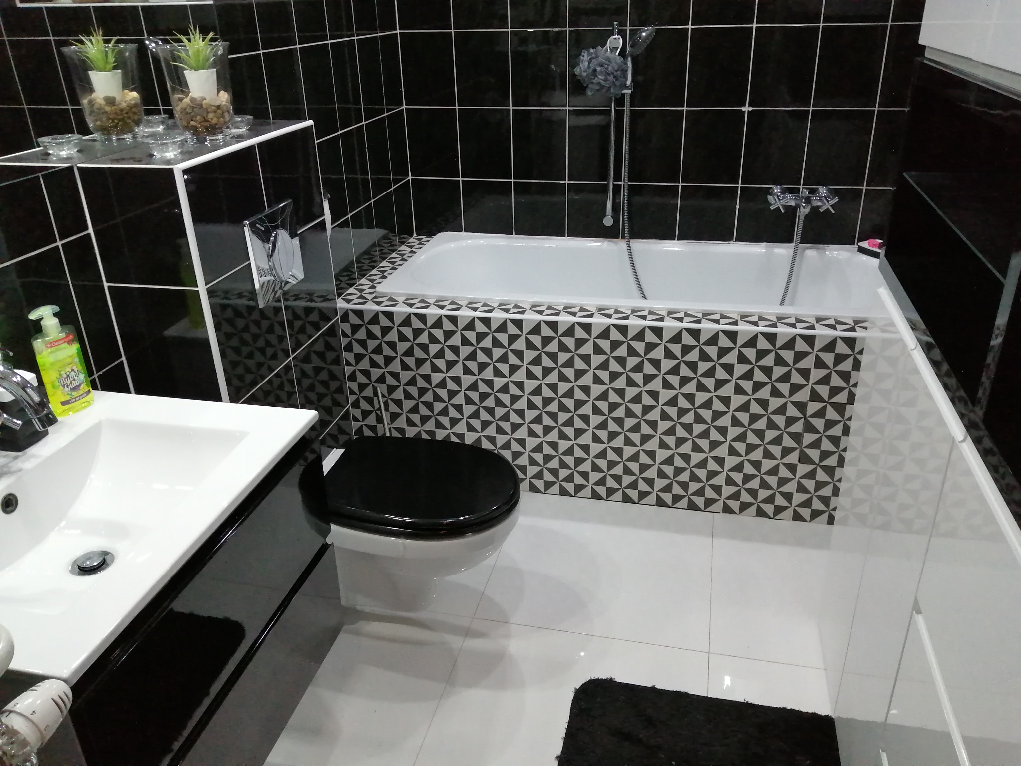 Nowoczesna łazienka w czarno-białej kolorystyce z wanną obudowaną w geometryczne wzory, białą umywalką z czarnym blatem, wiszącą toaletą z czarną deską oraz czarnymi płytkami na ścianach i białymi...