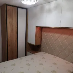 Sypialnia z zabudową meblową: szafa przesuwna z frontami drewnopodobnymi i białymi, szafki wiszące nad łóżkiem z podświetlanym zagłówkiem w romby.