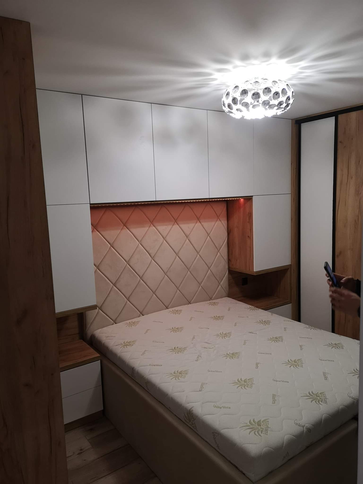 Nowoczesna sypialnia z łóżkiem, tapicerowanym zagłówkiem w romby podświetlonym od góry, szafkami nocnymi z szufladami, szafkami wiszącymi i szafą z przesuwnymi drzwiami w odcieniach drewna.