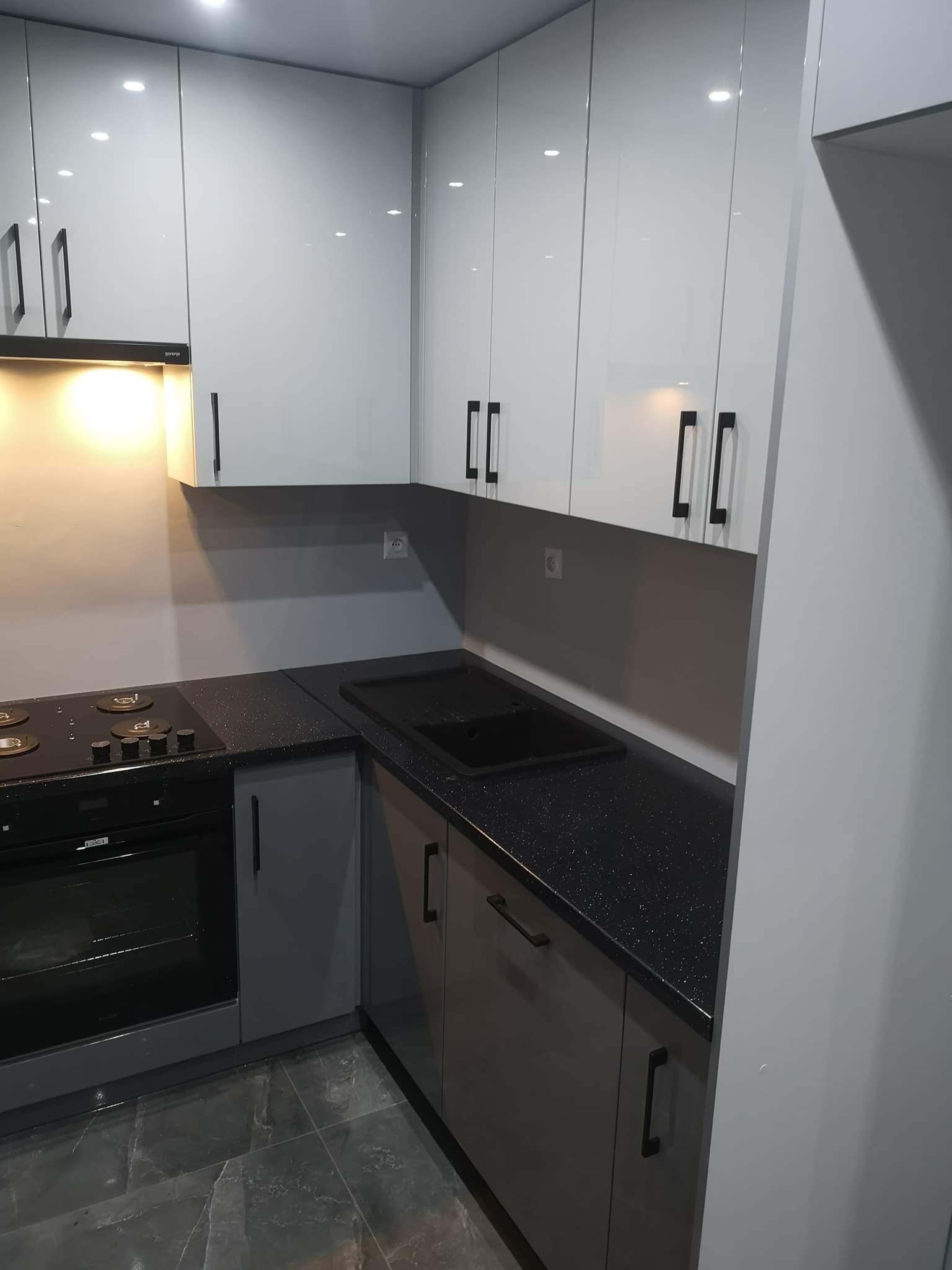 Nowoczesna kuchnia w odcieniach szarości i bieli z błyszczącymi frontami szafek i czarnym, połyskującym blatem z drobinkami brokatu, widoczny piekarnik i płyta grzewcza.