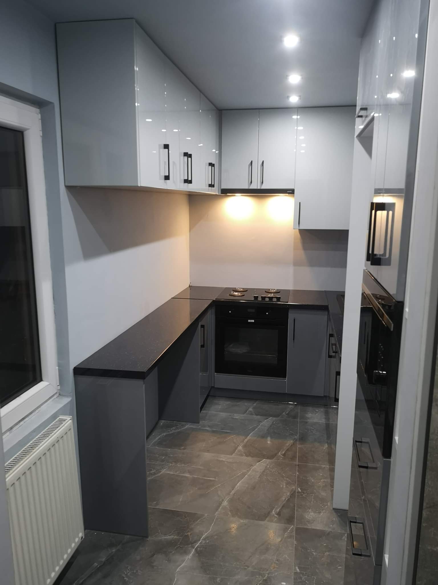 Nowoczesna, szara kuchnia z połyskiem, czarny blat z brokatem i marmurowa podłoga, widziana z perspektywy wejścia.