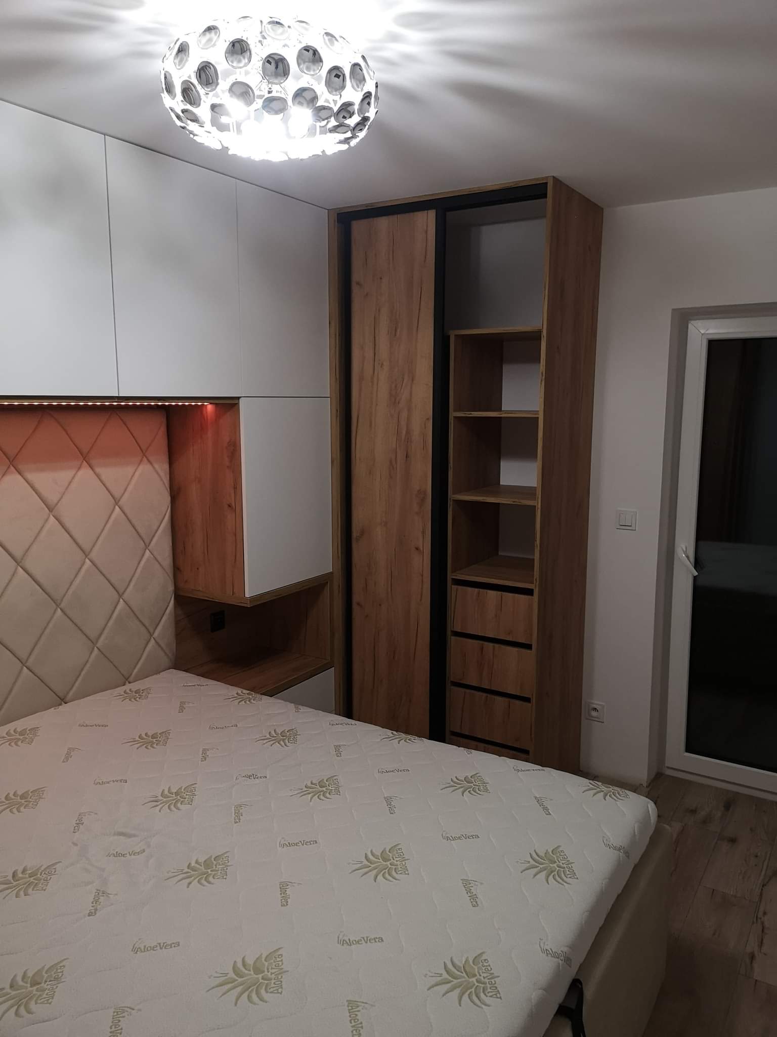 Nowoczesna sypialnia z łóżkiem, tapicerowanym zagłówkiem w romby, szafą przesuwną w kolorze drewna i białymi szafkami wiszącymi, oświetlona lampą sufitową