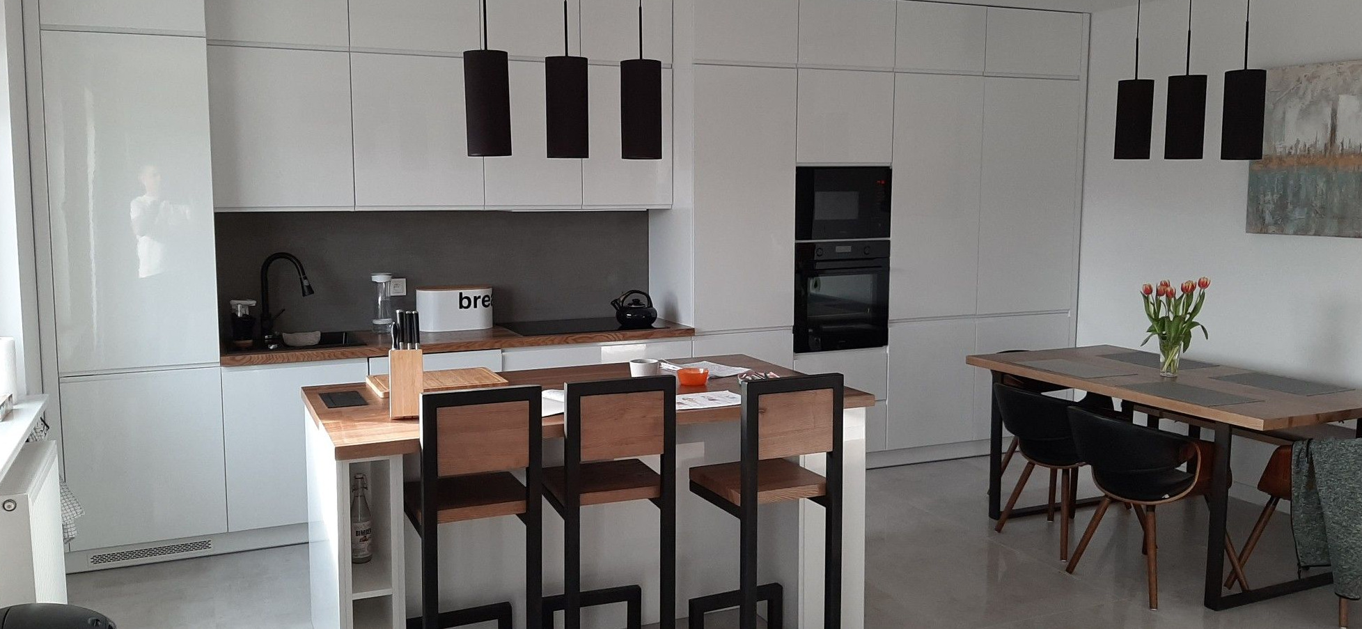 Nowoczesna kuchnia z białymi, lakierowanymi szafkami, wyspą z drewnianym blatem i hokerami z czarną metalową konstrukcją, oświetlona cylindrycznymi lampami wiszącymi; w tle stół z bukietem tulipanów.