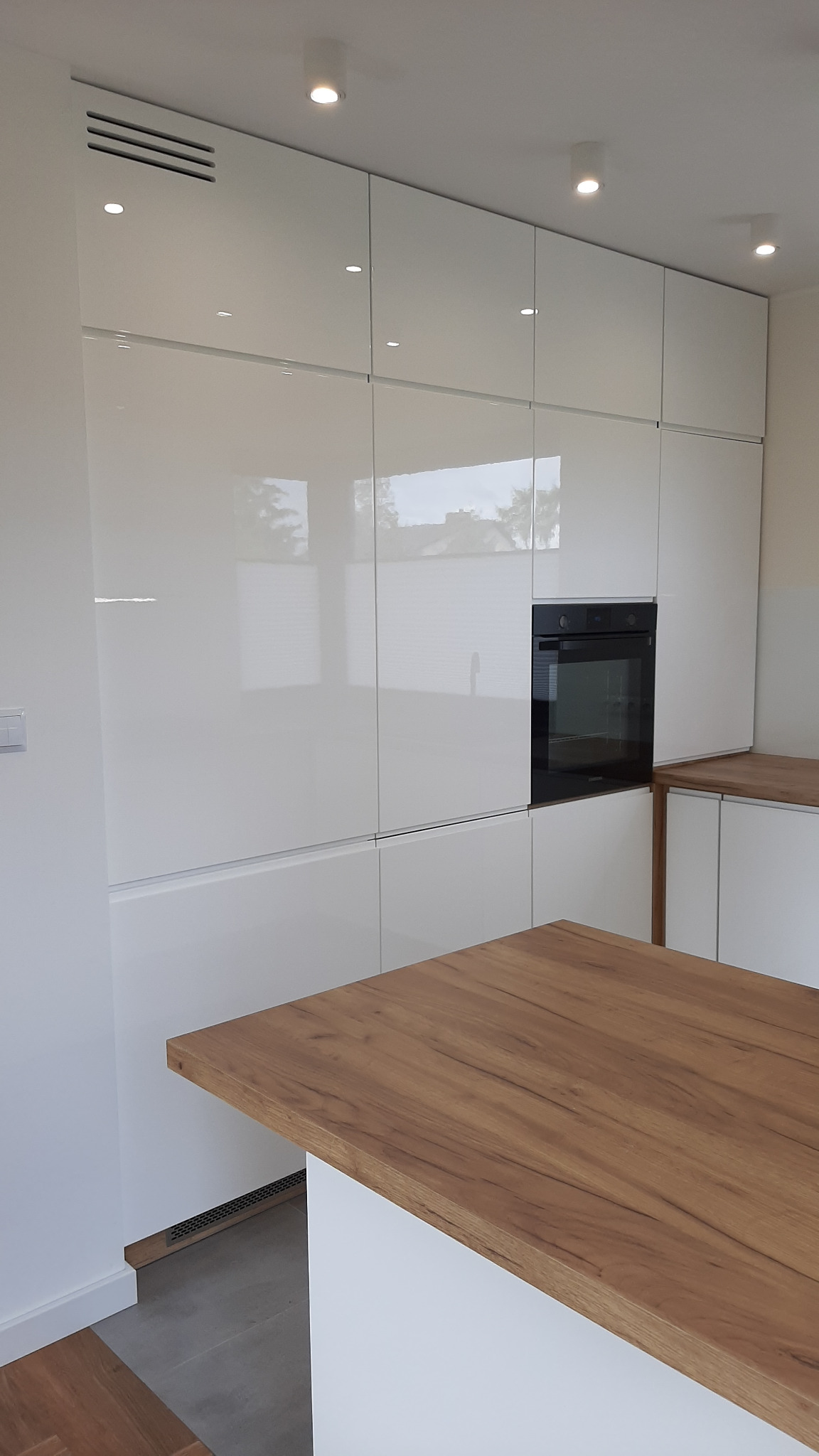 Biała zabudowa kuchenna z lakierowanymi frontami, wbudowanym piekarnikiem i blatem w kolorze drewna, widoczne detale wykończenia i oświetlenie punktowe.