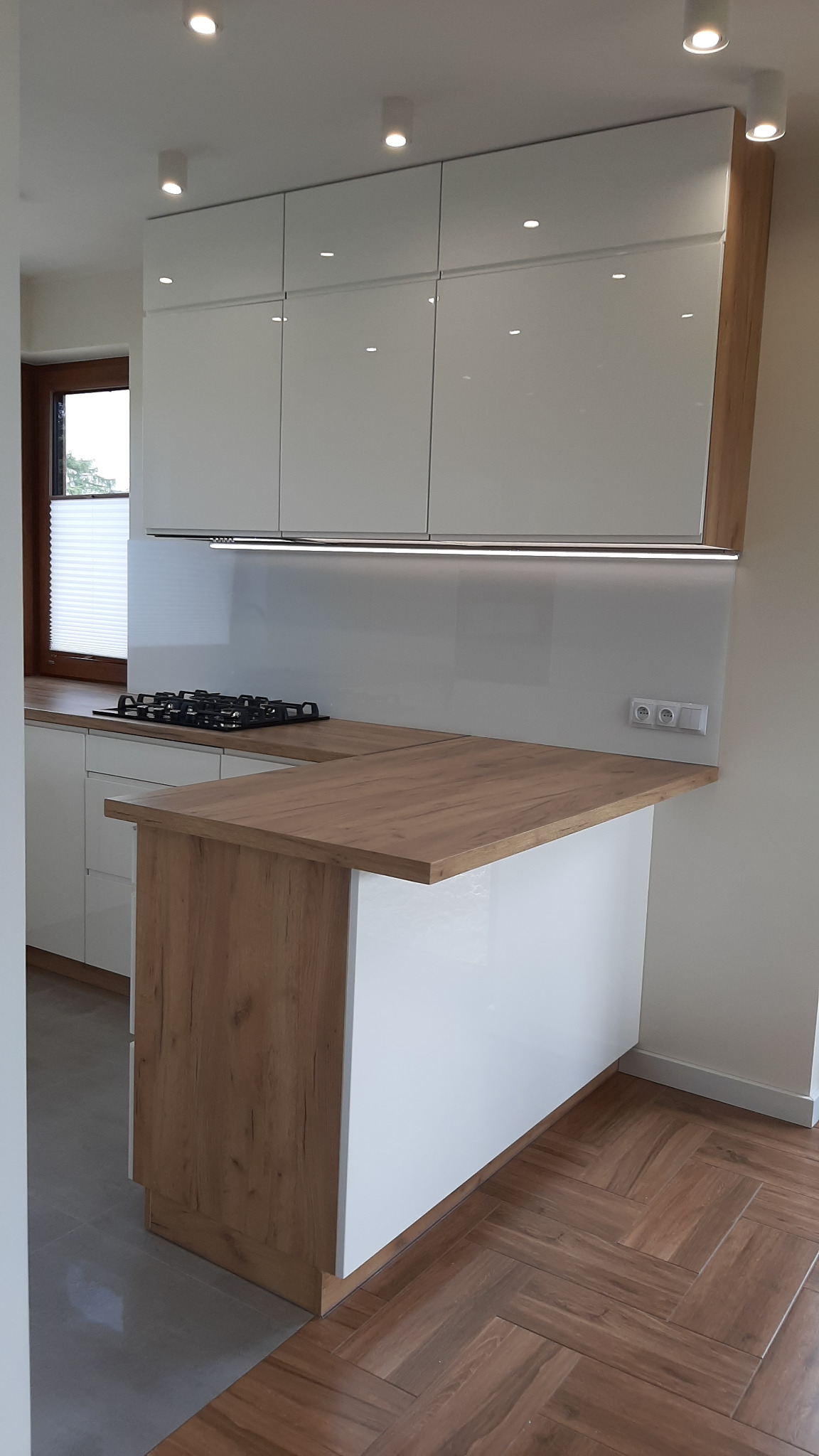 Nowoczesna kuchnia z białymi, lakierowanymi szafkami górnymi, drewnianym blatem wyspowym i podświetleniem LED pod szafkami, widok na płytę gazową i parkiet w jodełkę.