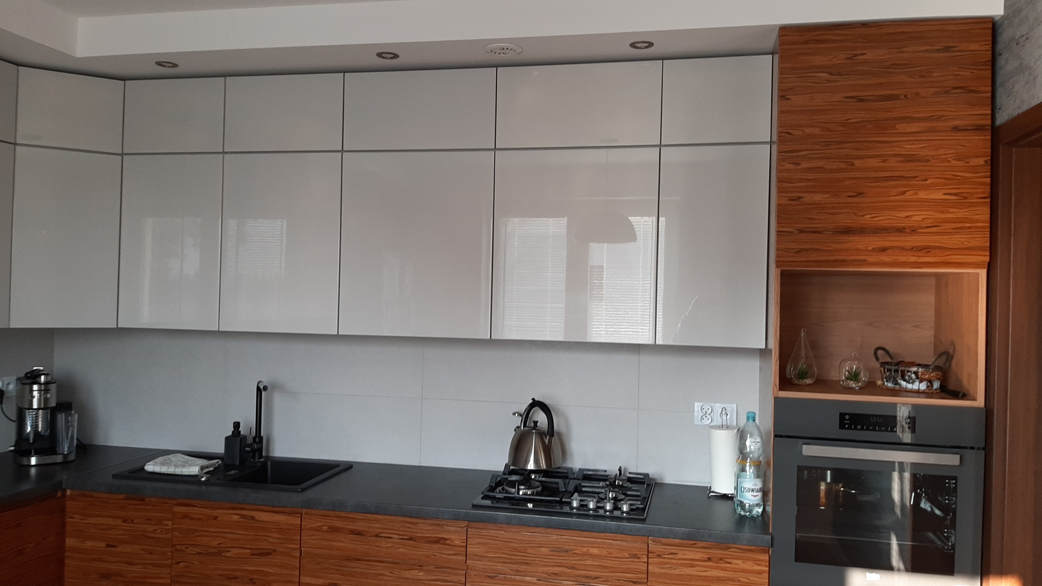 Nowoczesna kuchnia z białymi, błyszczącymi szafkami górnymi i drewnopodobnymi szafkami dolnymi w Niedrzwicy Dużej, z czarnym zlewem i płytą grzewczą.