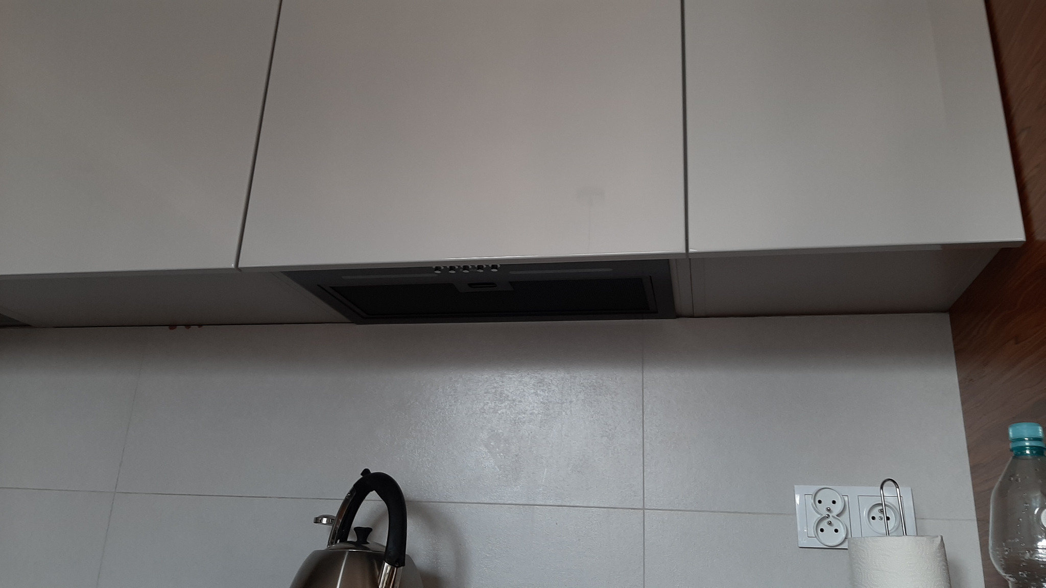 Białe szafki kuchenne z szarym okapem, srebrny czajnik i białe płytki na ścianie.