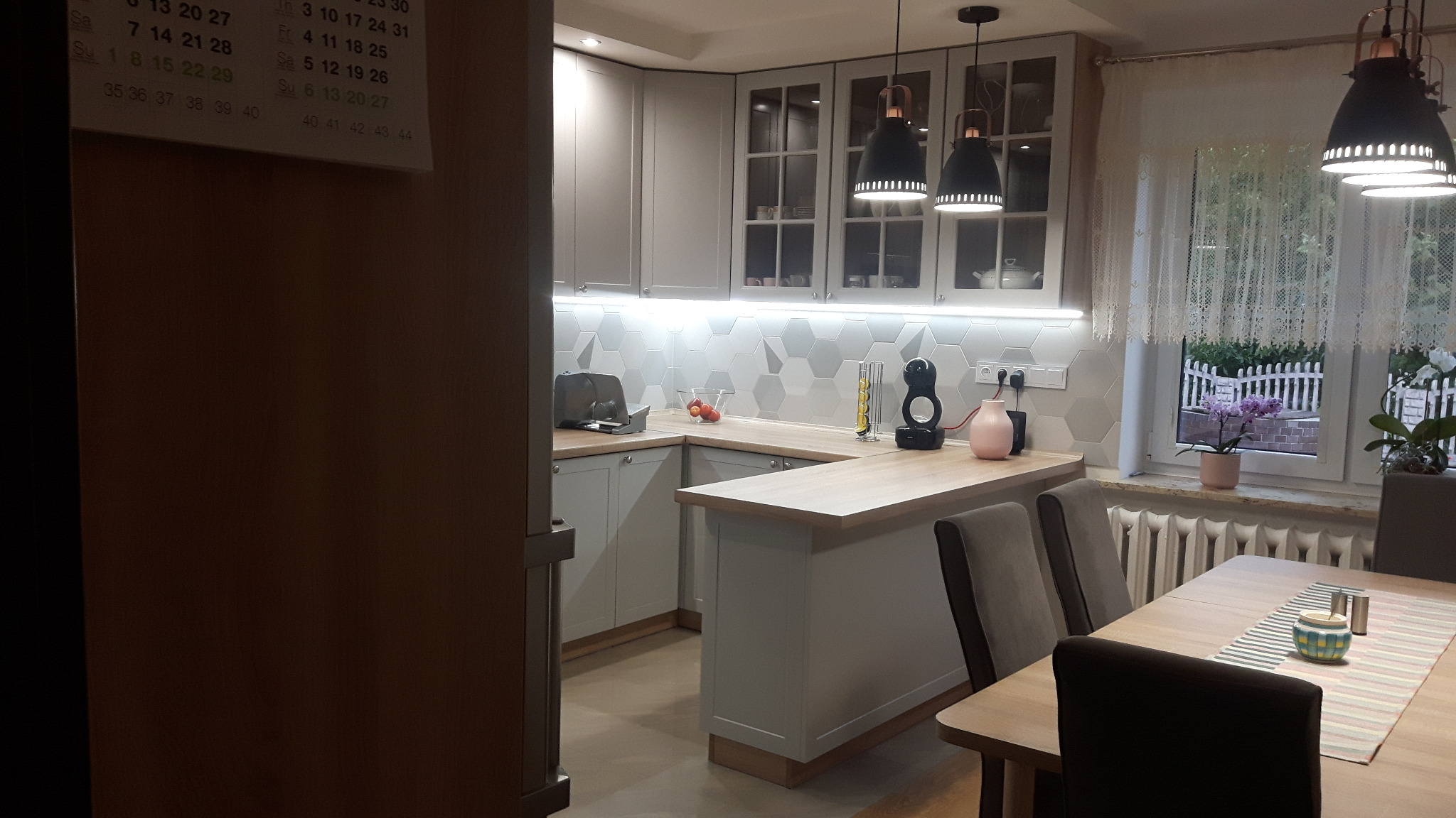 Jasnoszara zabudowa kuchenna z drewnianym blatem i barkiem, heksagonalne płytki na ścianie, wiszące lampy z miedzianymi detalami, widok na okno z kwiatami i grzejnikiem.