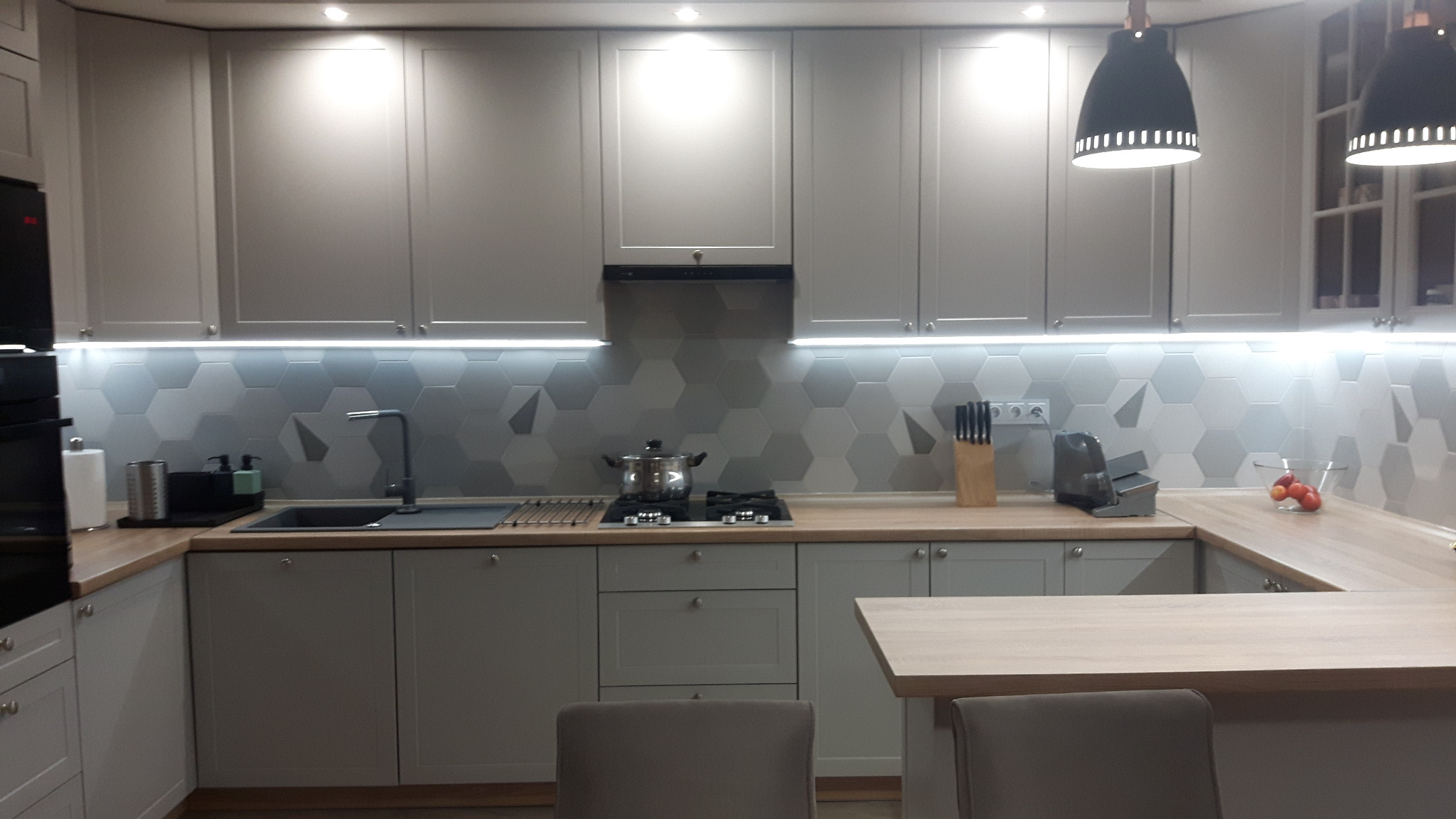 Szara kuchnia z heksagonalnymi płytkami na ścianie, oświetleniem LED pod szafkami i wiszącymi lampami w stylu skandynawskim.