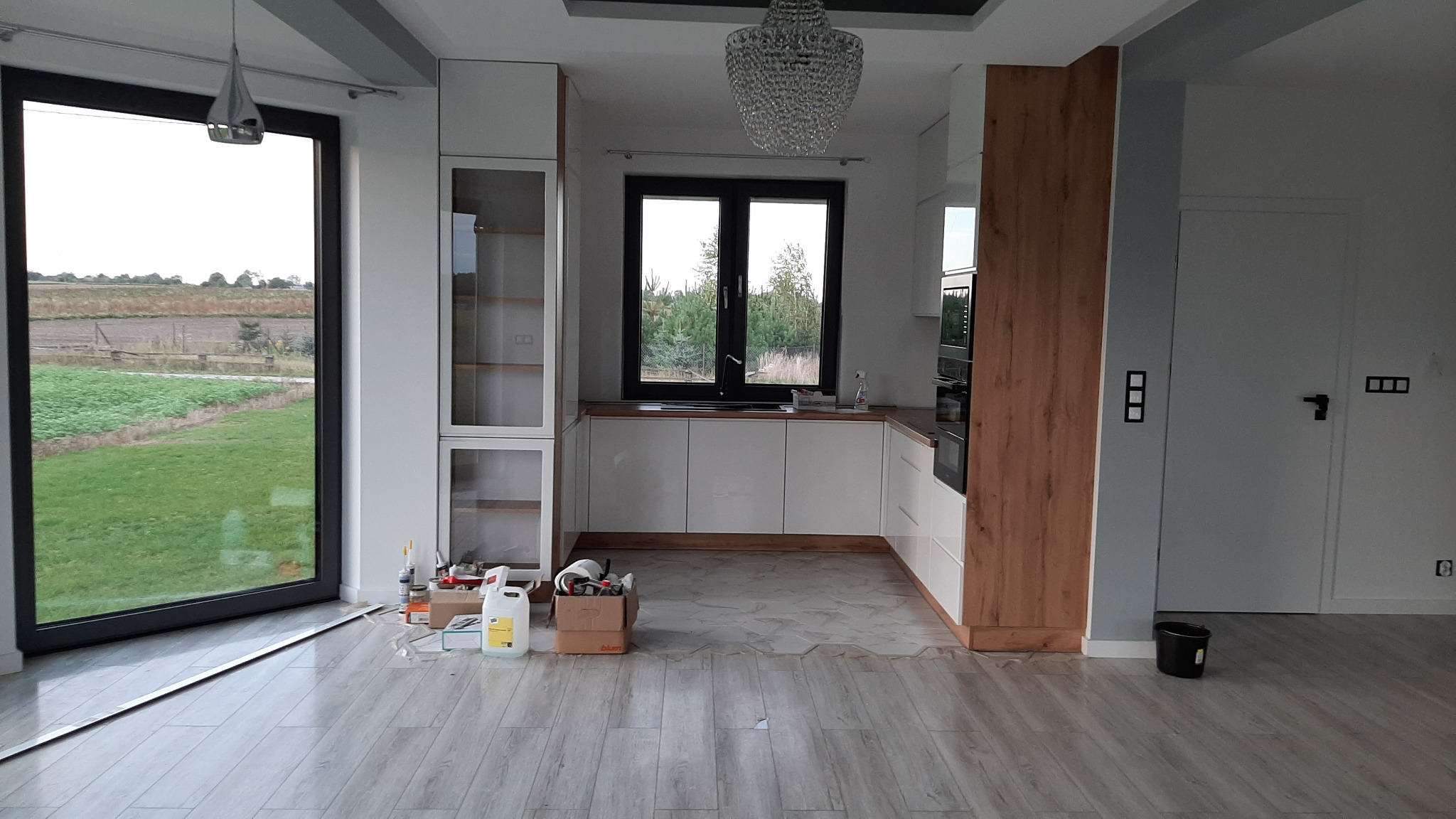 Jasna kuchnia z białymi szafkami i drewnianym blatem pod oknem z widokiem na zieleń, w trakcie montażu, z widocznymi narzędziami i materiałami na podłodze, nad nią kryształowy żyrandol.