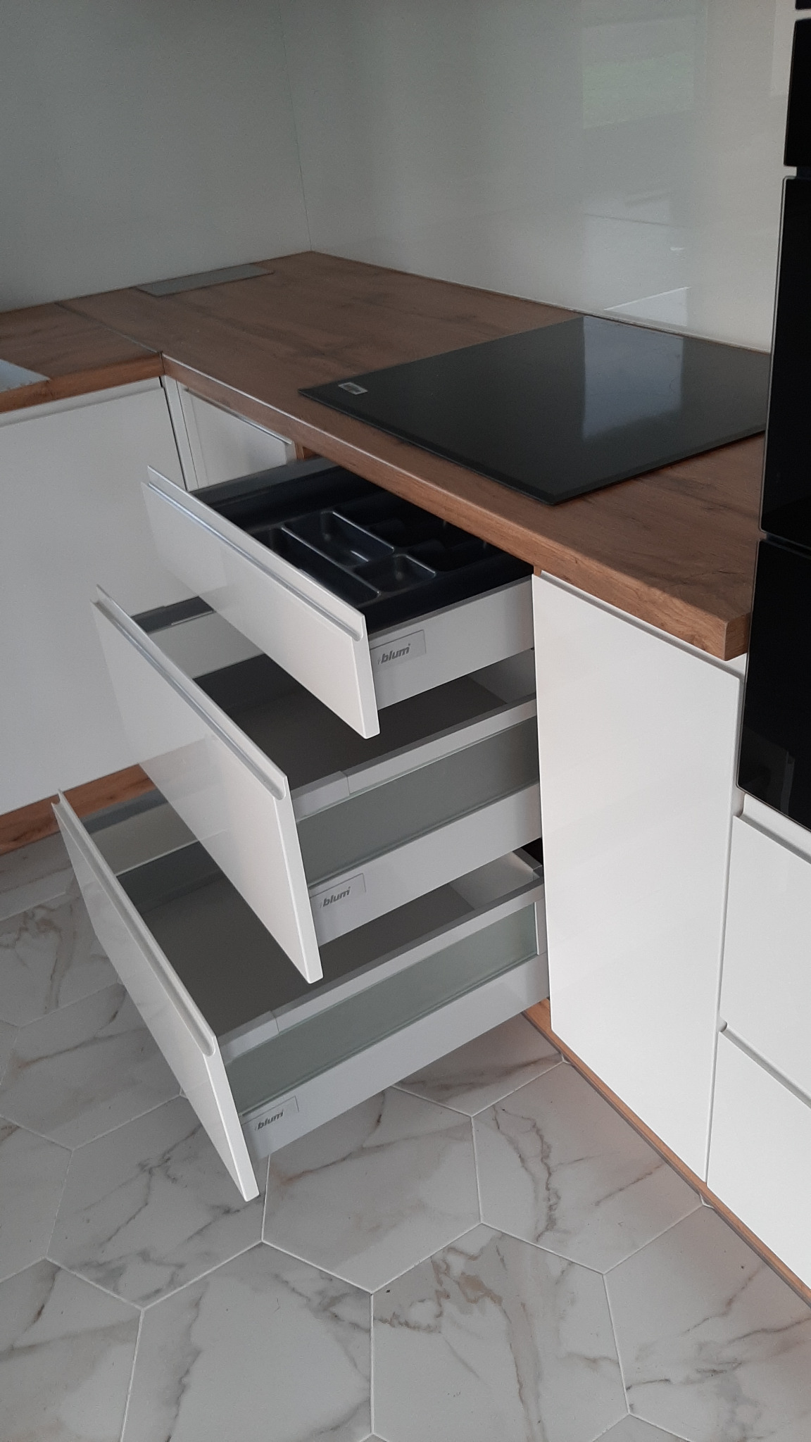 Nowoczesna kuchnia z białymi frontami i drewnianym blatem, widoczne trzy wysuwane szuflady z systemem Blum, górna z organizerem na sztućce, marmurowe płytki podłogowe w kształcie heksagonów.