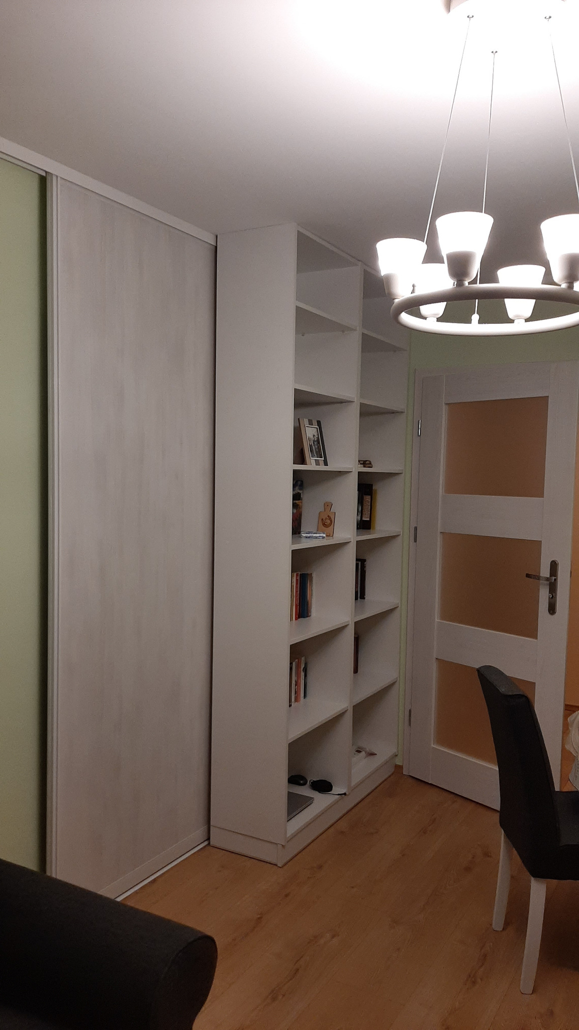 Biała zabudowa meblowa na wymiar: szafa przesuwna z jasnego drewna i regał z półkami, wypełniony książkami i dekoracjami, oświetlony nowoczesnym żyrandolem.