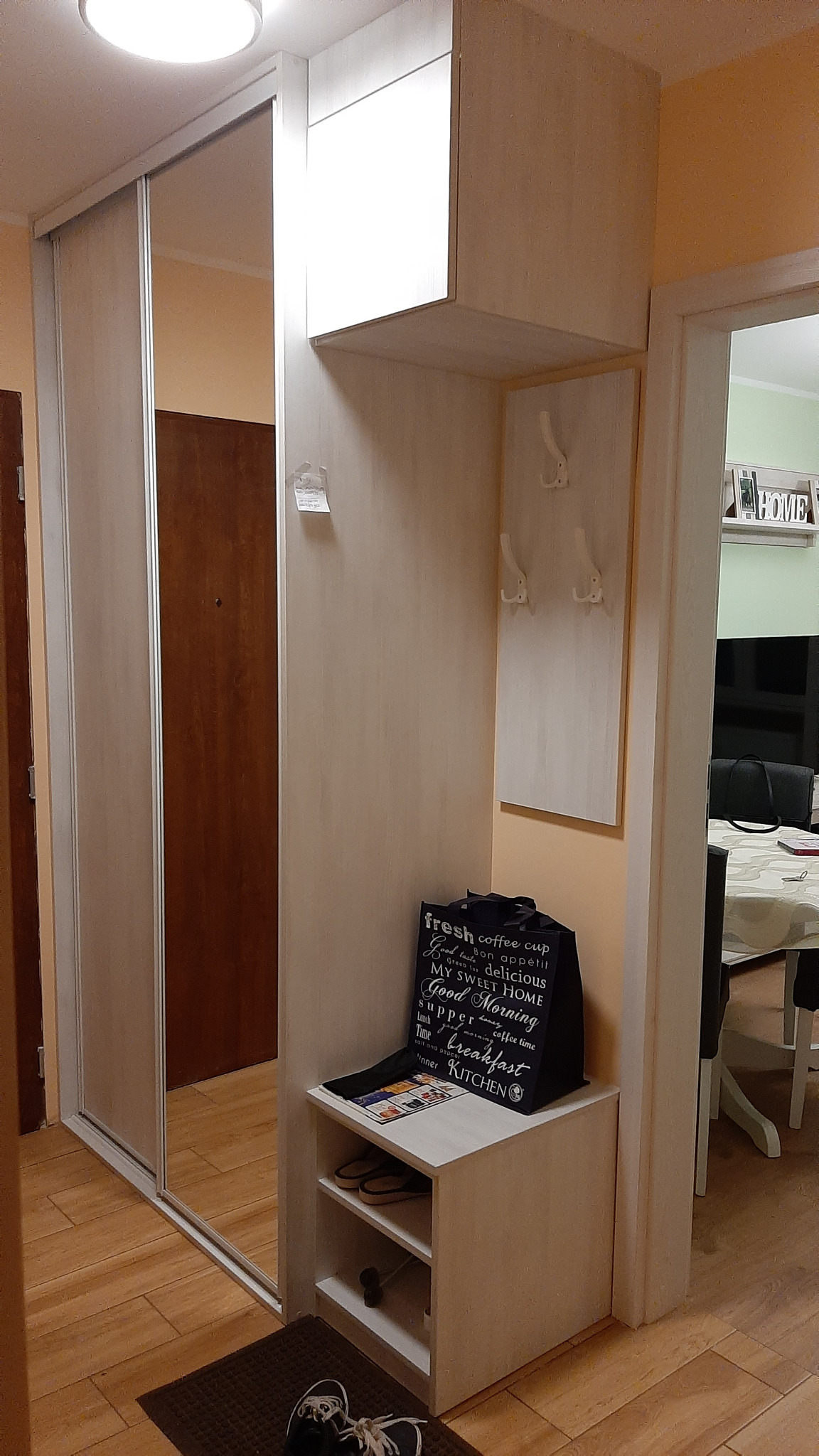 Przedpokój z szafą przesuwną z lustrem i wnęką z wieszakami oraz półkami na buty, na których leży czarna torba z napisem 'fresh coffee cup'.