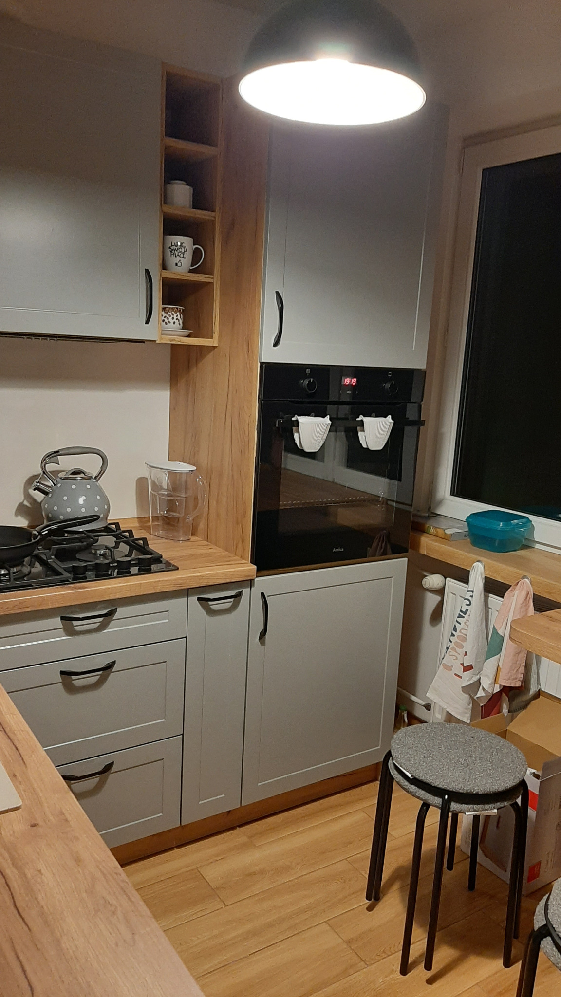 Szara zabudowa kuchenna z drewnianymi blatami, piekarnikiem i otwartą półką z filiżankami, oświetlona wiszącą lampą.