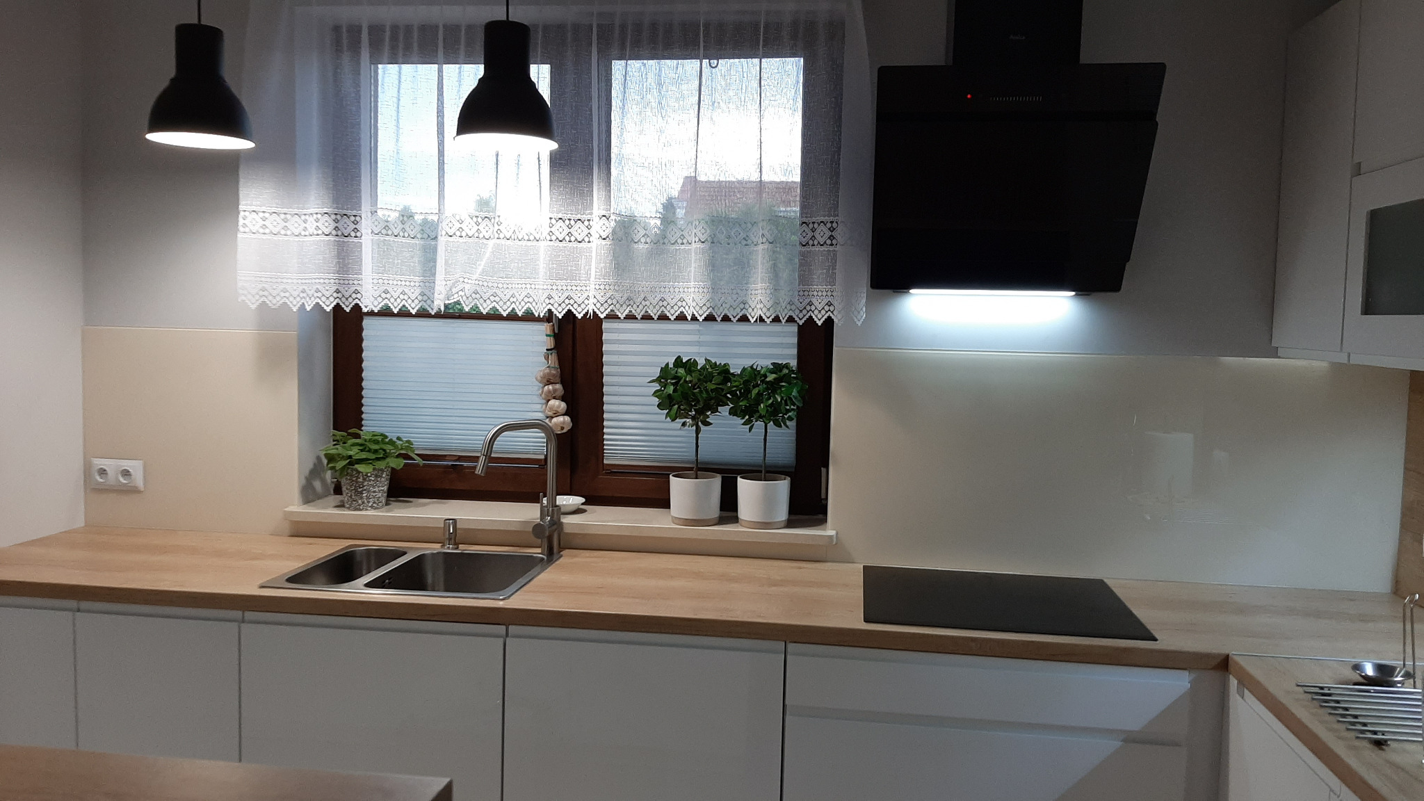 Nowoczesna kuchnia z białymi szafkami, drewnianym blatem, dwukomorowym zlewem i oknem z roletami plisowanymi, ozdobionym czosnkiem i doniczkami z drzewkami.