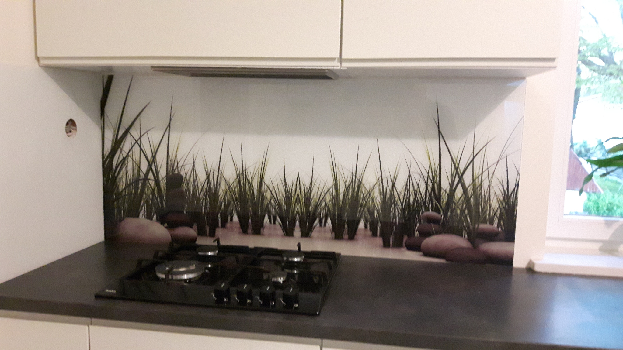 Szklany panel ścienny w kuchni z motywem trawy i kamieni zen, zamontowany nad czarną płytą gazową, widoczne białe szafki wiszące i fragment okna.