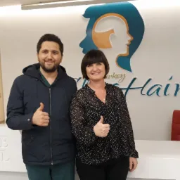 Mężczyzna i kobieta uśmiechnięci, pokazujący kciuki w górę na tle logo kliniki przeszczepu włosów 'Turkey Hair' w recepcji szpitala.