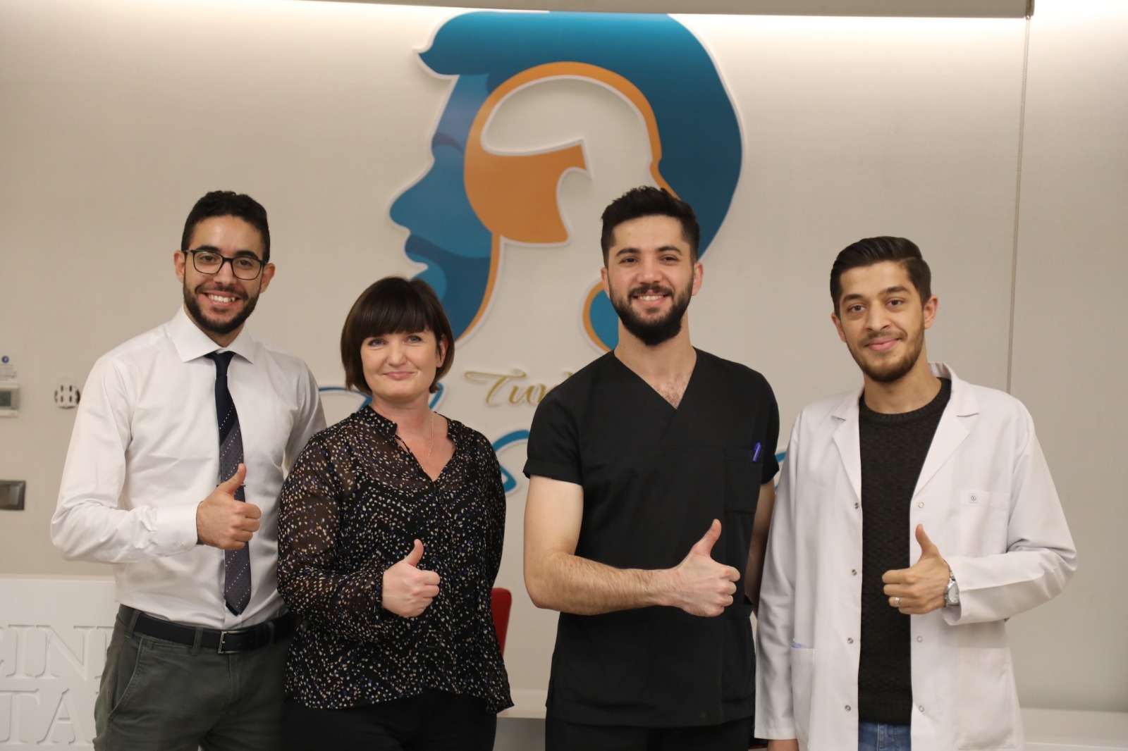 Zespół medyczny kliniki medycyny estetycznej, czterech uśmiechniętych pracowników z uniesionymi kciukami na tle logo firmy w kształcie profilu twarzy.