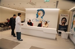 Wnętrze kliniki Class Hair z recepcją, przy której stoi lekarz w białym fartuchu, a za nią siedzą pacjenci; w tle widoczny baner reklamowy z wizerunkiem mężczyzny.