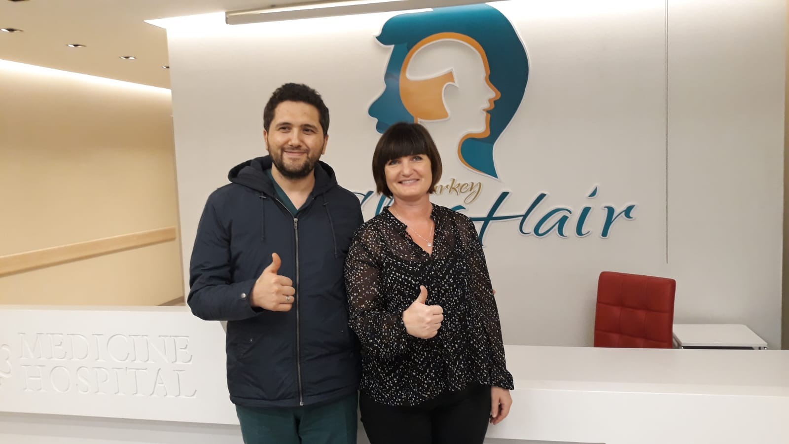 Mężczyzna i kobieta uśmiechnięci, pokazujący kciuki w górę na tle logo kliniki przeszczepu włosów 'Turkey Hair' w recepcji szpitala.