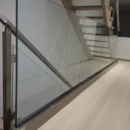 Szklana balustrada przy schodach z drewnianymi stopniami, mocowana do podłogi i bocznej ściany, z metalowymi słupkami i listwami wykończeniowymi.