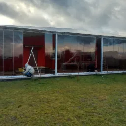 Montażysta w szarej kurtce instaluje szklane panele przesuwne w konstrukcji altany z czerwonym wnętrzem, drabina oparta o ścianę, pochmurne niebo w tle.