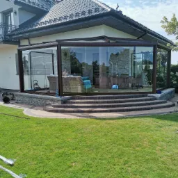 Szklana zabudowa tarasu z brązowymi ramami, widok z zewnątrz na schody prowadzące do wnętrza, częściowo rozpakowane elementy konstrukcyjne na trawniku.