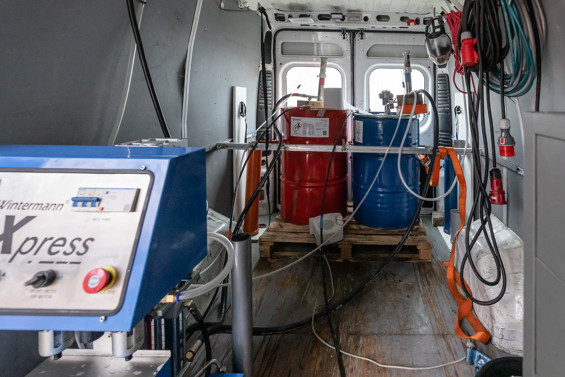 Wnętrze furgonetki z zamontowanym urządzeniem Wintermann Xpress, dwoma beczkami (czerwona i niebieska) z substancjami chemicznymi na palecie, przewodami i kablami, prawdopodobnie do aplikacji...