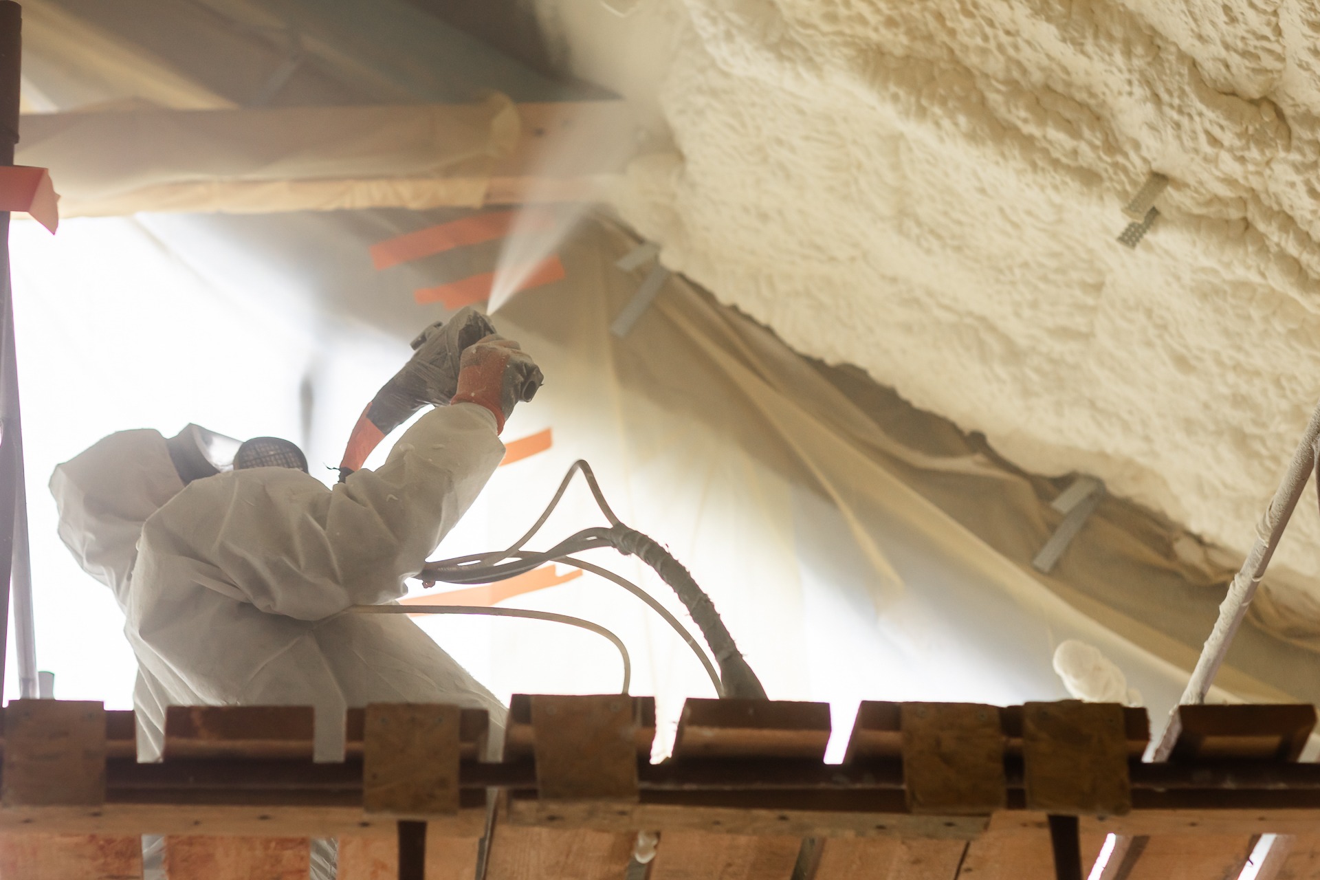 Pracownik w kombinezonie ochronnym, z bliska, natryskuje pianę izolacyjną na poddasze; widoczna struktura piany i drewniane elementy konstrukcyjne dachu.