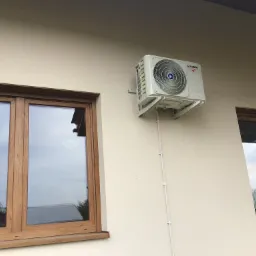 Zewnętrzna jednostka klimatyzacji Kaisai zamontowana na ścianie budynku pomiędzy dwoma oknami z drewnianymi ramami, widoczna biała listwa maskująca przewody.