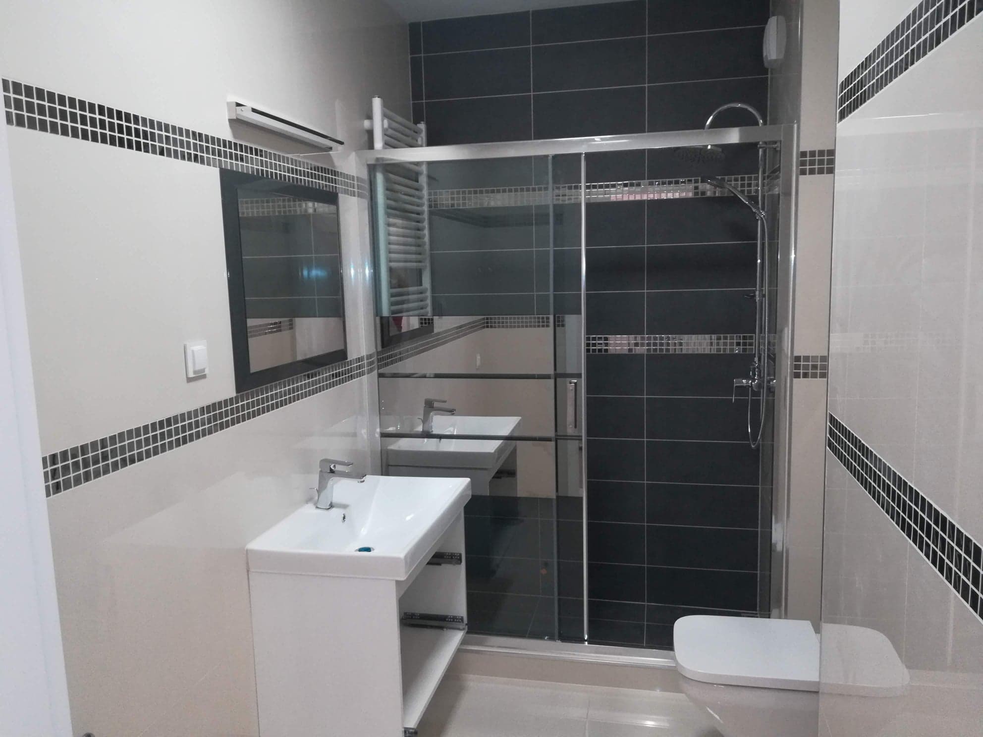 Nowoczesna łazienka z czarno-białymi płytkami, kabiną prysznicową ze szklanymi drzwiami przesuwnymi i białą umywalką z szafką, ozdobiona pasem mozaiki.