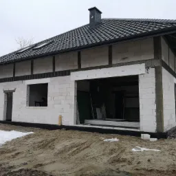 Budowa domu jednorodzinnego w Rudniku nad Sanem w stanie surowym otwartym: białe bloczki, czarna dachówka, widoczne otwory okienne i garażowe. Teren wokół domu częściowo pokryty śniegiem i błotem.