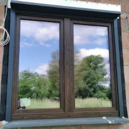 Zamontowane okno dwuskrzydłowe z brązowymi ramami i szarą roletą zewnętrzną, widoczne elementy montażowe i odbicie krajobrazu w szybach, surowy mur z cegły.