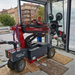 Montaż dużej szklanej tafli okiennej za pomocą specjalistycznego robota z przyssawkami, widoczni pracownicy w trakcie instalacji, budynek mieszkalny w tle.