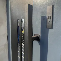 Nowoczesne, grafitowe drzwi wejściowe z podłużnymi przeszkleniami, masywną klamką i zamkiem, zamontowane w surowym otoczeniu.