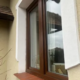 Okno PCV z brązową ramą, parapet wyłożony czerwoną cegłą, widok z zewnątrz, biała rama okienna z widocznymi pęknięciami.