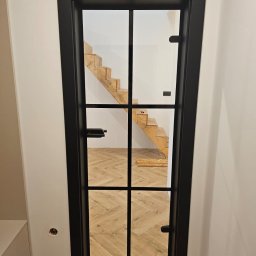 FHU PROFIL DOM Jan Konior - Czarne, przeszklone drzwi wewnętrzne z kratownicą, widok na drewniane schody i parkiet ułożony w jodełkę. Minimalistyczny design, jasne ściany.