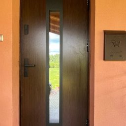 FHU PROFIL DOM Jan Konior - Ciemnobrązowe drzwi wejściowe z wąskim, pionowym przeszkleniem, zamontowane w pomarańczowej ścianie budynku. Widoczna skrzynka pocztowa i fragment podjazdu.