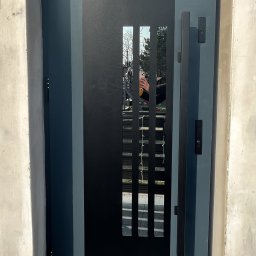 FHU PROFIL DOM Jan Konior - Nowoczesne, grafitowe drzwi zewnętrzne z podłużnym przeszkleniem w metalowej ramie, widoczne na tle surowego betonu. Minimalistyczny design z długim, pionowym uchwytem.