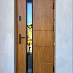 FHU PROFIL DOM Jan Konior - Nowoczesne drzwi wejściowe z drewnopodobnym panelem i pionowym przeszkleniem, czarne okucia, osadzone w surowym, jasnym murze.