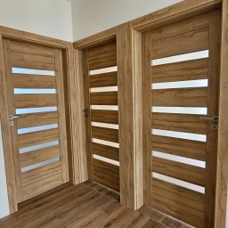 FHU PROFIL DOM Jan Konior - Trzy drewniane drzwi wewnętrzne z poziomymi przeszkleniami, zamontowane w jasnym pomieszczeniu z podłogą imitującą drewno. Widok z bliska, prezentujący detale wykończenia.