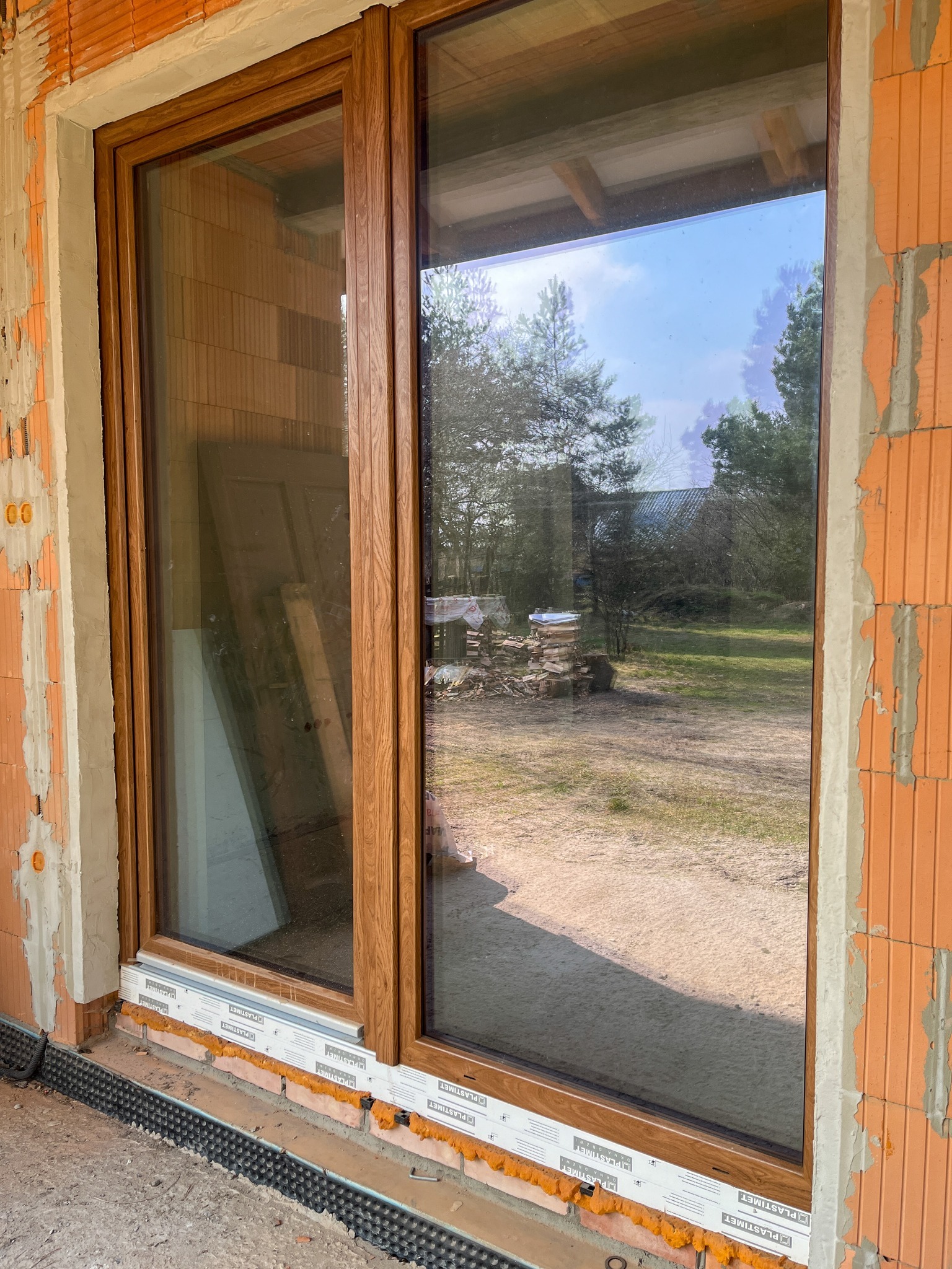 Montaż okna PCV z drewnopodobną ramą, widoczne cegły i materiały budowlane, odblask w szybie ukazuje drzewa i budynek w tle.