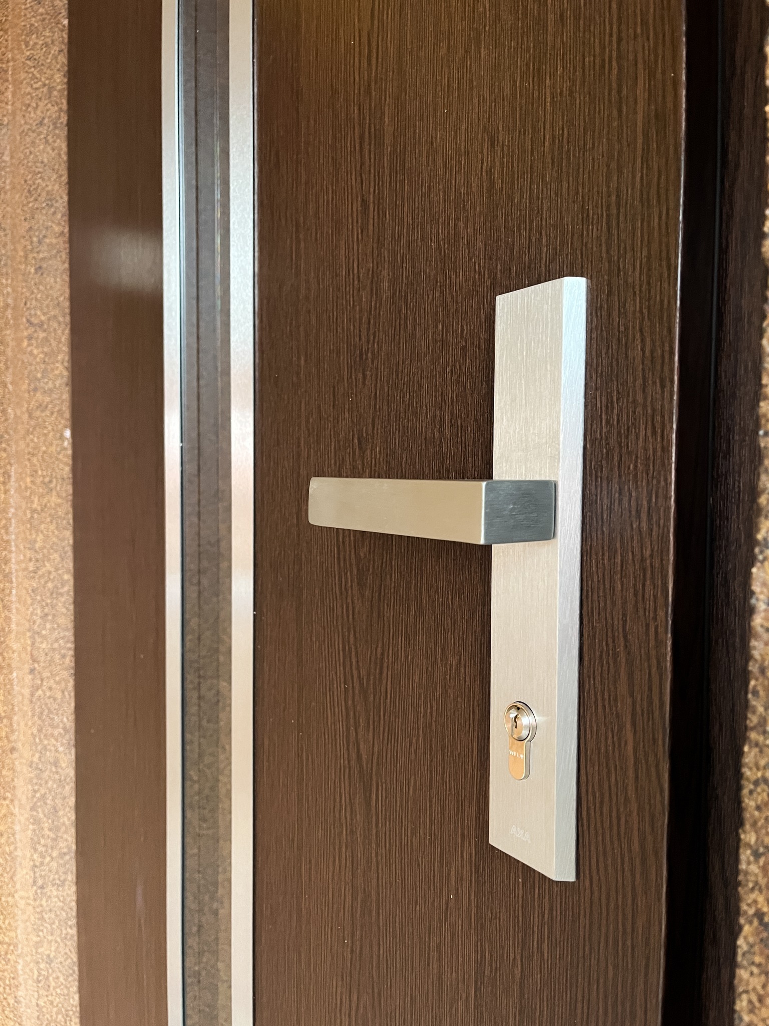 Brązowe drzwi z drewnopodobnym wykończeniem, z prostą, srebrną klamką i zamkiem, widoczne fragmenty ościeżnicy i ściany.