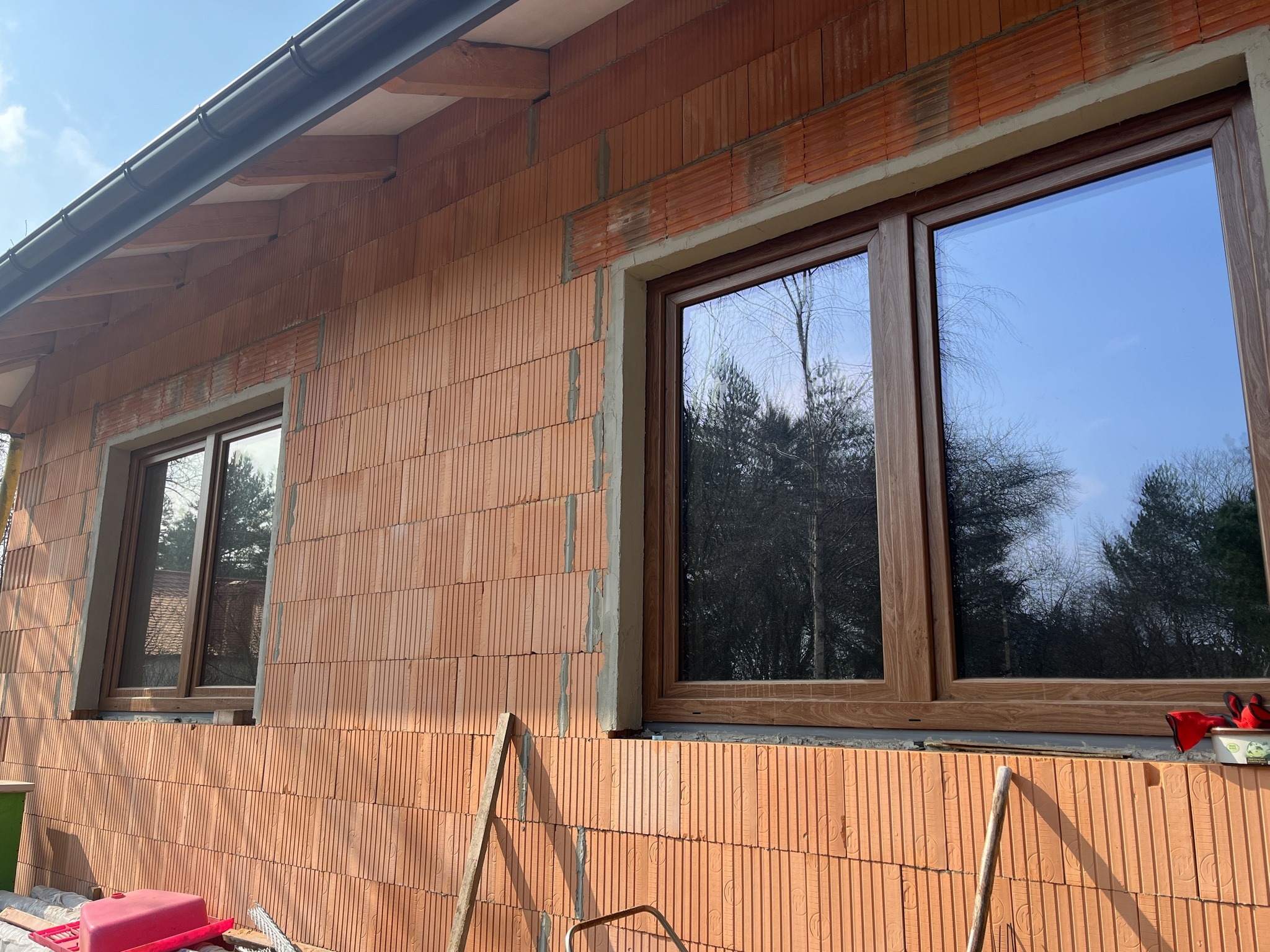 Dwa okna z brązowymi ramami zamontowane w surowej ścianie z czerwonej cegły, widoczne narzędzia i akcesoria budowlane, odbicie drzew w szybach.