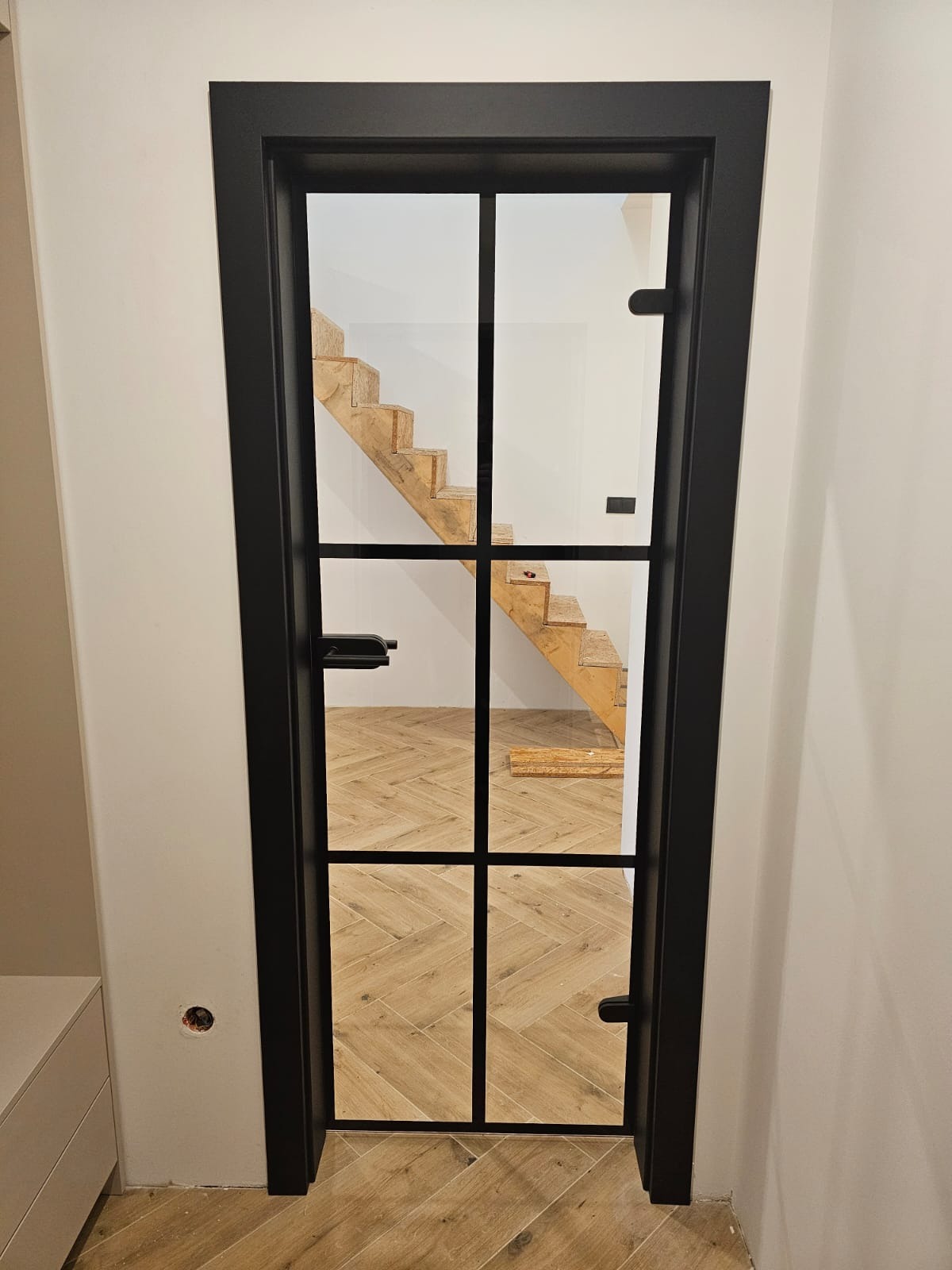 Czarne, przeszklone drzwi wewnętrzne z kratownicą, widok na drewniane schody i parkiet ułożony w jodełkę. Minimalistyczny design, jasne ściany.