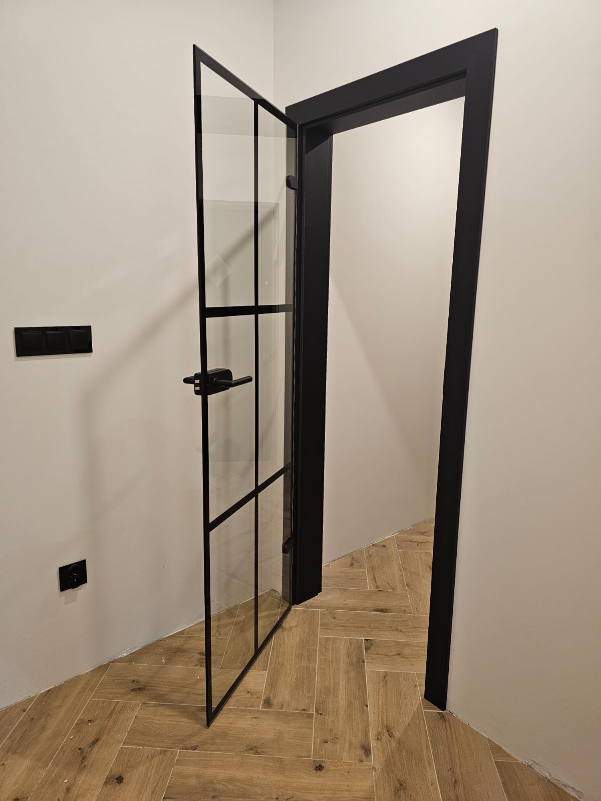 Czarne, loftowe drzwi szklane z metalową ramą, częściowo otwarte, ukazujące wnętrze. Widoczna klamka i fragment podłogi z drewnopodobnych płytek.