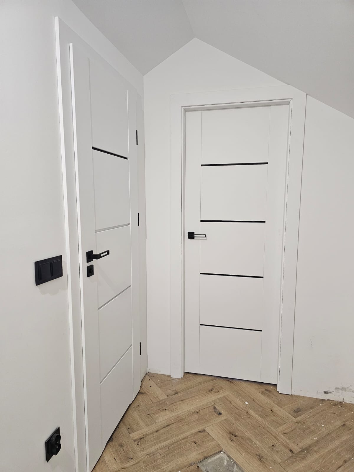 Dwie białe, nowoczesne drzwi wewnętrzne z czarnymi akcentami i klamkami, zamontowane w jasnym wnętrzu z parkietem ułożonym w jodełkę.