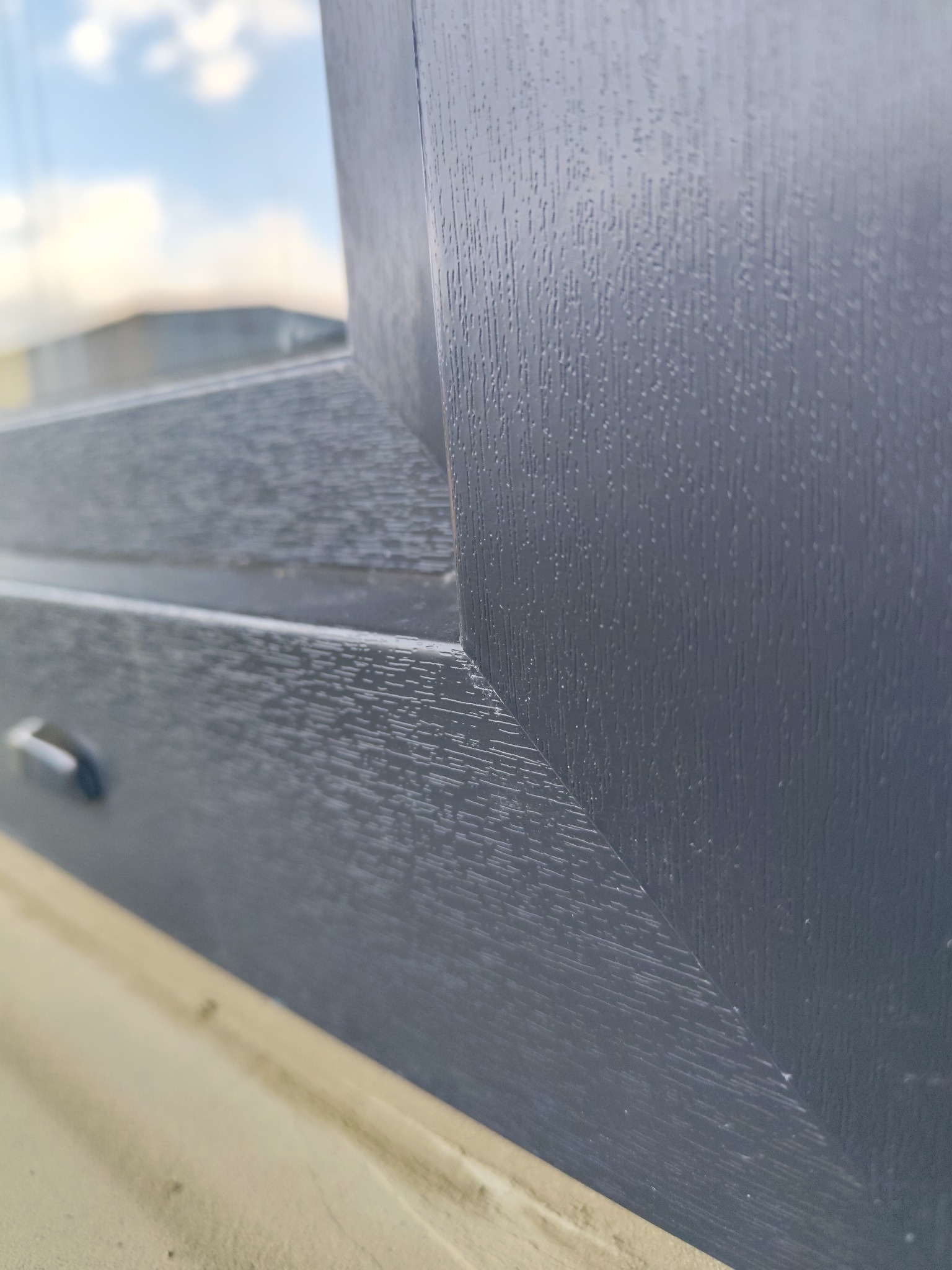 Szczegółowe zbliżenie na grafitową ramę okienną z wyraźną fakturą drewna, widoczne niebo w tle. Ujęcie z niskiego kąta podkreśla detale wykończenia.